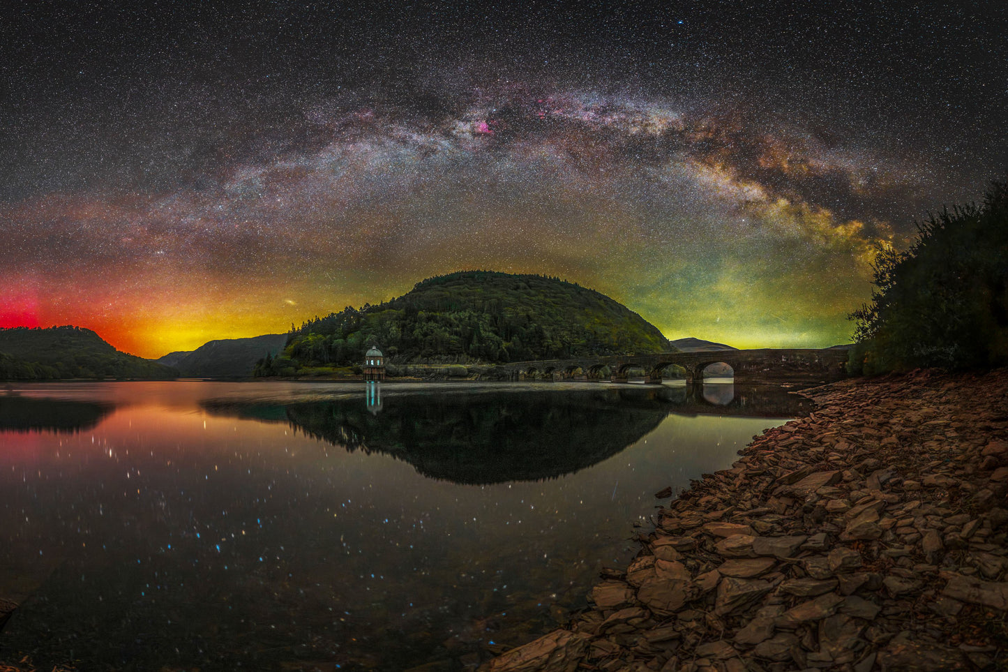 Panorama composite of the Spring Milky Way arch and Aurora Borealis reflected in the still waters of Garreg Ddu reservoir, Elan Valley, Wales, with the Victorian waterworks building and dam visible at the water's edge.