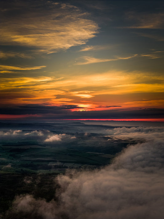 Drone view above the mist on the North York Moors at summer solstice sunset, with vivid orange and yellow skies glowing over a sea of cloud