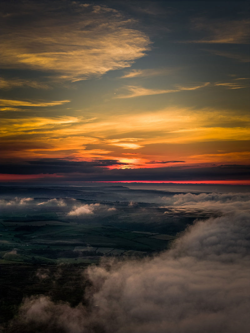 Drone view above the mist on the North York Moors at summer solstice sunset, with vivid orange and yellow skies glowing over a sea of cloud