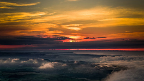 Drone view above the mist on the North York Moors at summer solstice sunset, with vivid orange and yellow skies glowing over a sea of cloud
