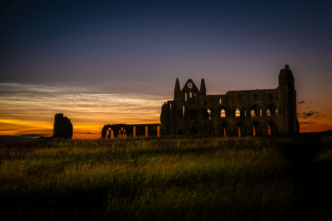 Noctilucent clouds glowing above the silhouette of Whitby Abbey ruins against twilight skies, with long grass in the foreground illuminated by a nearby security light