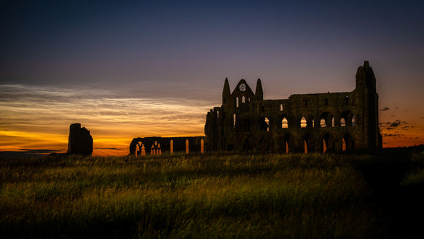 Noctilucent clouds glowing above the silhouette of Whitby Abbey ruins against twilight skies, with long grass in the foreground illuminated by a nearby security light